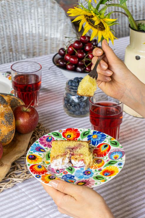 A woman's hand holding a bite of cake on a fork over a folk dessert plate.